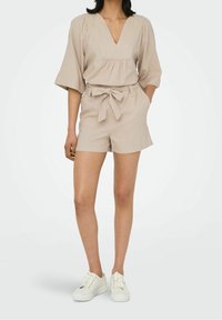 Combinaison short beige avec un col en V, manches bouffantes, ceinture nouée en tissu à la taille, finition texturée et coupe décontractée.