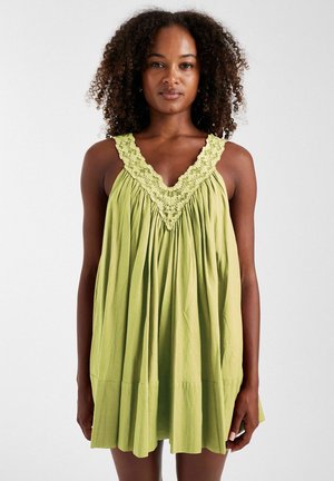 Femme aux cheveux bouclés portant une robe verte ample sans manches avec un bord en dentelle au niveau du décolleté, debout devant un fond uni.
