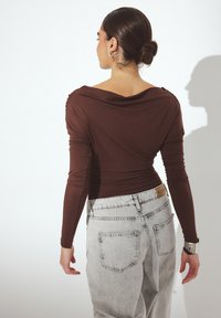 Brown long-sleeve top with a draped neckline paired with light gray denim jeans featuring back pockets and a light wash finish.