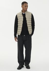 Mees, kes seisab käed taskus, kantud beežis vooderdatud vestis, mustas pika varrukaga särgis, lõdvades mustades pükstes ja mustades kingades valgel taustal.