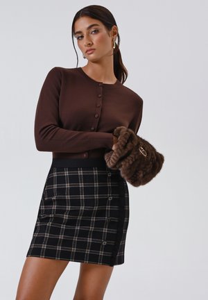 Donna che indossa un cardigan marrone con bottoni, una gonna scozzese nera e marrone con bottoni, mentre tiene in mano una borsa in finta pelliccia marrone, su uno sfondo neutro.