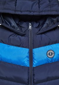 Giubbotto imbottito navy con striscia orizzontale blu, chiusura con zip, cappuccio e patch circolare. Tessuto liscio e texture trapuntata.