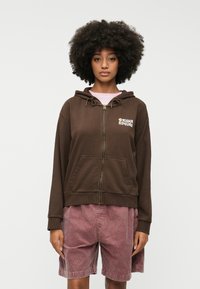 Καφέ φούτερ με φερμουάρ και μπροστινό λογότυπο, διαθέτει τσέπη καγκουρό και κορδόνια στην κουκούλα. Συνδυασμένο με ροζ καμπαρντίνες σορτς.