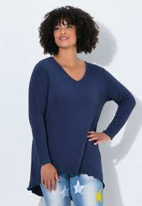 Kobieta z kręconymi włosami, nosząca granatowy długi rękaw V-neck oraz niebieskie dżinsy w białe i żółte gwiazdy, pozująca z ręką na biodrze.