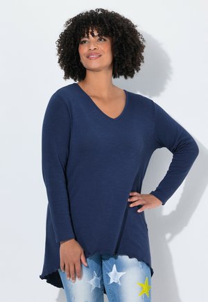Femme aux cheveux bouclés portant un haut à manches longues bleu marine en V et un jean bleu avec des motifs d'étoiles blanches et jaunes, posant avec une main sur la hanche.
