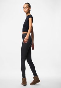 Μαύρη εφαρμοστή κοντομάνικη μπλούζα συνδυασμένη με σκούρους τζιν και καφέ σουέτ μποτάκια μέχρι τον αστράγαλο. Μοντέλο που στέκεται στο πλάι σε λευκό φόντο.