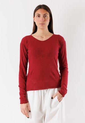 Donna con lunghi capelli scuri che indossa una maglietta rossa a maniche lunghe e pantaloni bianchi, in piedi con una mano in tasca davanti a uno sfondo uniforme.