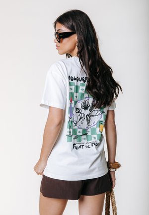 Femme aux longs cheveux foncés, portant de grandes lunettes de soleil noires, un t-shirt blanc à motif coquillage, un short marron et tenant un sac tressé.