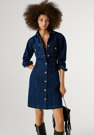 Robe chemise en denim bleu foncé, dotée de boutons sur le devant, de poches poitrine et de manches retroussées. Texture lisse, longueur au-dessus du genou.