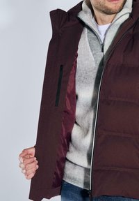 Bordeauxrote Steppjacke mit Reißverschlusstasche, gefüttert mit dunkelrotem Stoff, getragen über einem hellgrauen Pullover mit gerippter Struktur.