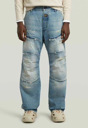 Light blue denim pants with distressed texture, featuring patchwork design, visible seams, and rips. Paired with tan slip-on shoes.