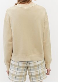 Sudadera beige con cuello redondo y hombros caídos, hecha de material suave, combinada con pantalones cortos de cuadros en verde claro y beige.