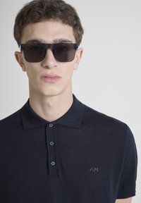Navyblauw polo shirt met korte mouwen heeft een kraag, drie knopen en een subtiel logo. Model draagt grote zwarte zonnebril.