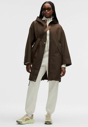 Woman with braided hair wearing dark brown hooded coat, white zip-up top, white jogger pants, beige sneakers, and black sunglasses, standing.