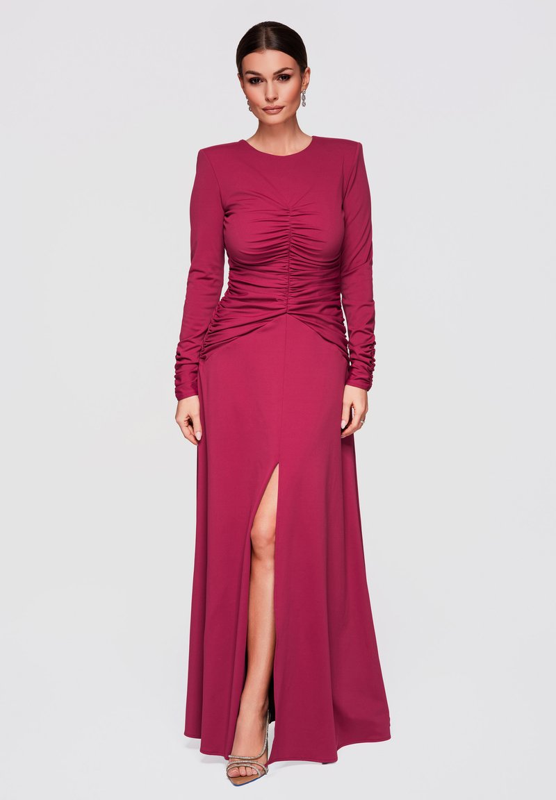 Femme portant une robe longue à manches longues froncée de couleur rose foncé avec une fente haute à l'avant sur la cuisse et des talons argentés à brides, debout devant un fond uni.
