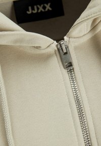 Hoodie beige avec une fermeture éclair en métal, présentant une texture côtelée et un col étiqueté avec un logo. Gros plan mettant en avant les détails du tissu et de la quincaillerie.