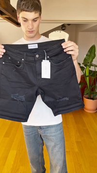 Calções de ganga pretos com bainhas desfiadas, múltiplos bolsos e uma etiqueta presa. A parte da frente apresenta detalhes desgastados. A modelo segura os calções.