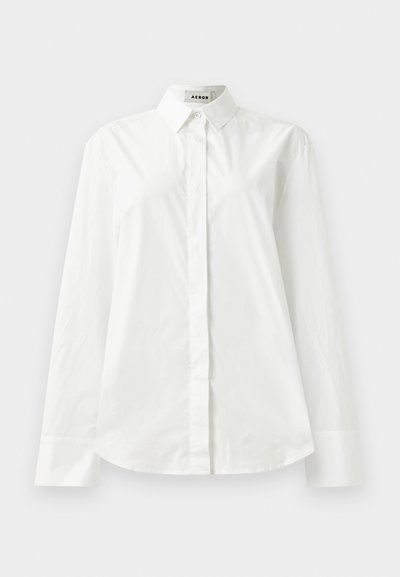 Camicia bianca in cotone a maniche lunghe con colletto a punta, chiusura anteriore con bottoni e orlo arrotondato. Tessuto liscio, vestibilità regolare e senza fantasie.