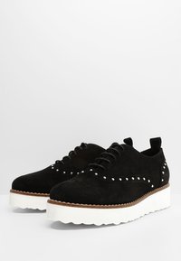 Zapatos de ante negros con cordones y suela gruesa blanca, que presentan tachuelas plateadas a lo largo del costado y un detalle de media suela cosido en marrón.