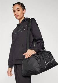 Bolsa de deporte negra tipo duffle con múltiples bolsillos con cremallera, tejido texturizado, asas reforzadas y una correa de hombro desmontable. Diseño liviano y duradero.