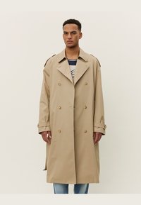 Gabardina beige con diseño doble botonadura, gran cuello y hombreras. Presenta acentos de botones y un corte a la altura de la rodilla.