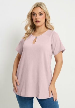 Blonde Frau trägt texturiertes, hellviolettes Kurzarm-Top mit Schlitz am Ausschnitt und blaue Jeans, posiert mit Händen seitlich.