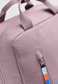 Pinkfarbene wasserdichte Tasche aus strukturiertem Stoff, mit seitlichem Reißverschlusselement und bunten Stoffakzenten. Wassertropfen auf der Oberfläche.