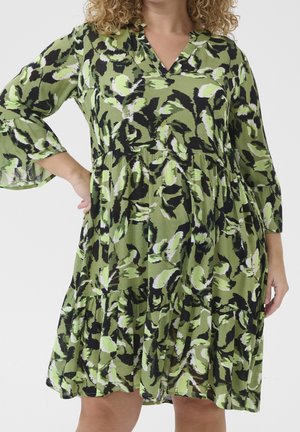 Woman wearing a green, black, and white abstract floral dress with three-quarter sleeves and a V-neck, standing with one hand on her hip.