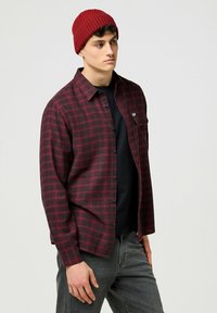 Camicia in flanella a quadri rossi e neri con chiusura a bottoni e tasca sul petto, indossata sopra una maglietta nera, abbinata a un berretto di lana rosso.