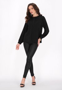 Blusa nera a maniche lunghe con dettagli plissettati, abbinata a pantaloni skinny in ecopelle neri e sandali con tacco alto neri. Tessuto liscio.