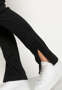 Černé strečové kalhoty s bočními rozparky u kotníků, spárované s bílými teniskami s hladkým koženým povrchem a šněrováním.