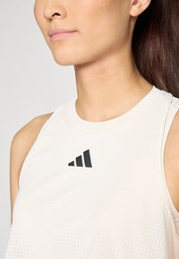 Débardeur de sport beige clair avec un col rond, arborant un logo noir et un motif texturé. Le matériau semble respirant et lisse.