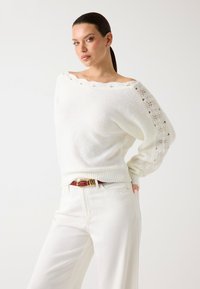 Pull en tricot blanc avec un design décolleté, motif de tricot ouvert sur les manches, ourlet côtelé, porté avec un pantalon blanc et une ceinture rouge.