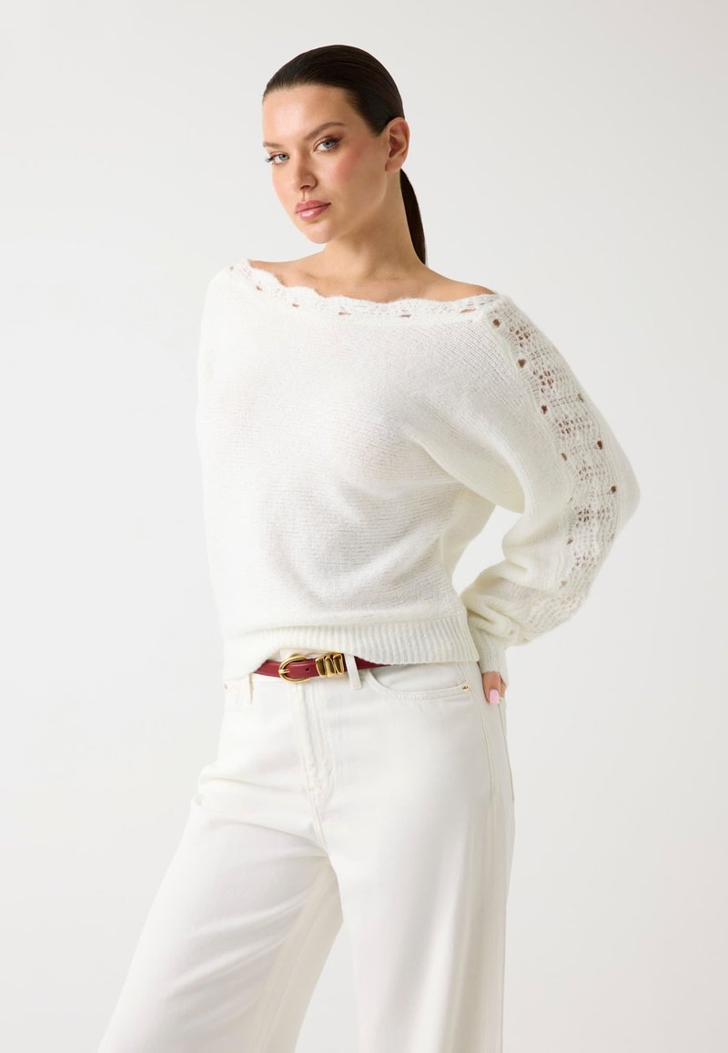 Pull en tricot blanc avec un design décolleté, motif de tricot ouvert sur les manches, ourlet côtelé, porté avec un pantalon blanc et une ceinture rouge.