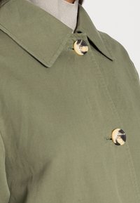 Olijfgroene jas met een spits kraag, voorzien van twee gevlekte knopen en fijne stikdetails op een zachte, gestructureerde stof.