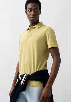 Νέος άνδρας που φοράει ανοιχτό πράσινο πόλο και ανοιχτό μπλε τζιν, με μαύρο μπουφάν δεμένο γύρω από τη μέση του, κοιτάζοντας πλάγια.