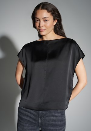 Femme aux longs cheveux noirs portant un haut ample en satin noir et un jean foncé, debout devant un fond gris clair uni.