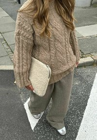 Bežani pulover s pletenim uzorkom i slobodnim krojem, uparen s sivim hlačama širokih nogavica. Drži bež clutch torbicu s teksturom. Naočite cipele su vidljive.