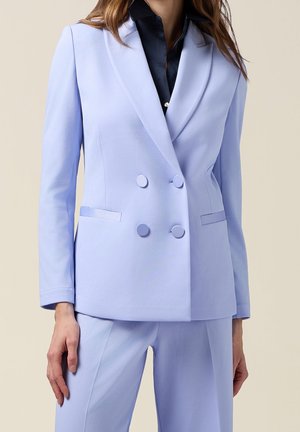 Femme portant un blazer bleu clair à double boutonnage avec un pantalon assorti et une chemise bleu foncé boutonnée en dessous.