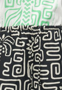 Shorts noirs avec des motifs abstraits en spirales et floraux blancs, bénéficiant d'une taille élastique avec un lien, associés à un haut blanc à impressions vertes.