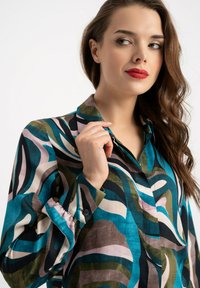 Kobieta z falowanymi brązowymi włosami i czerwonymi ustami, ubrana w bluzkę w wielokolorowy, abstrakcyjny wzór, trzymająca kołnierzyk palcami.