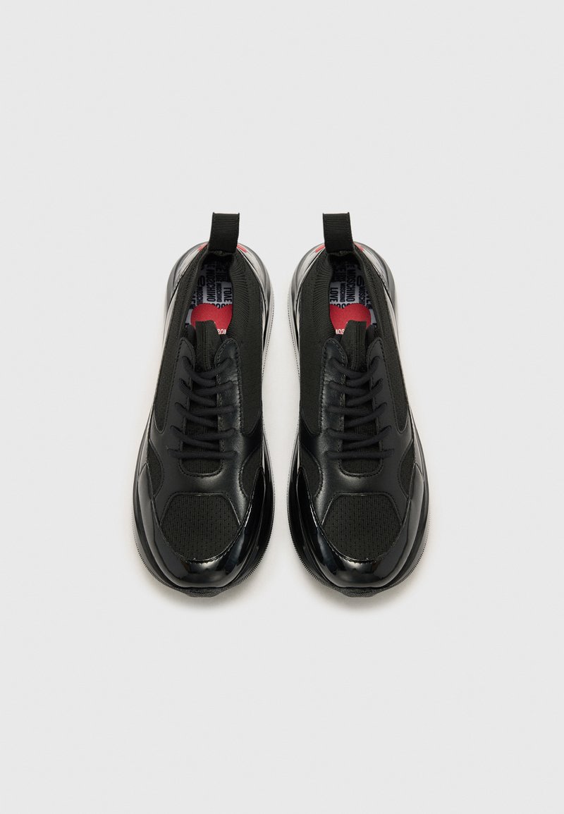 Scarpe da ginnastica nere con una finitura liscia e lucida, pannelli in rete testurizzati e un tassello per il sollevamento. Presentano un design con lacci e accenti rossi a contrasto.