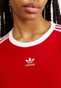 Červené tričko Adidas s bílým kulatým výstřihem, třemi bílými pruhy na rameni a malým bílým logem na hrudi.