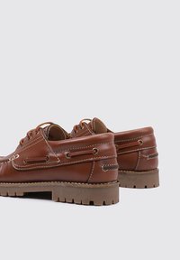 Zapatos de barco de cuero marrón con costuras elevadas, diseño de cordones y suelas de goma con patrones de tracción. Ojales metálicos visibles en la parte trasera.