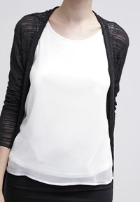 Žena v bílé bezrukávové halence pod průsvitným černým kardigánem s dlouhými rukávy, doplněná černou sukní na světlém pozadí.