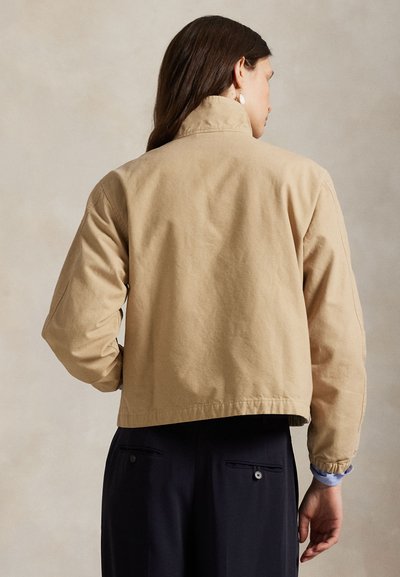 Kvinne sett bakfra, iført en beige kort jakke med lange ermer og høy krage over svarte bukser med baklommer som lukkes med knapper.