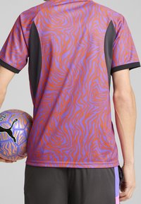 Fioletowo-pomarańczowy sportowy t-shirt w wzory z czarnymi akcentami, krótki rękaw i obcisły krój, trzymany przez osobę obok wzorowanej piłki nożnej.