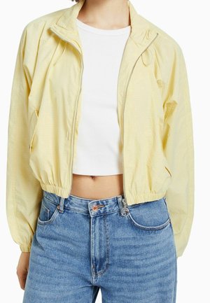 Osoba ubrana w jasnofiltrujący żółty wiatrówkę na zamek, biały krótki prążkowany top i niebieskie dżinsy z wysokim stanem, pokazana od szyi do bioder.