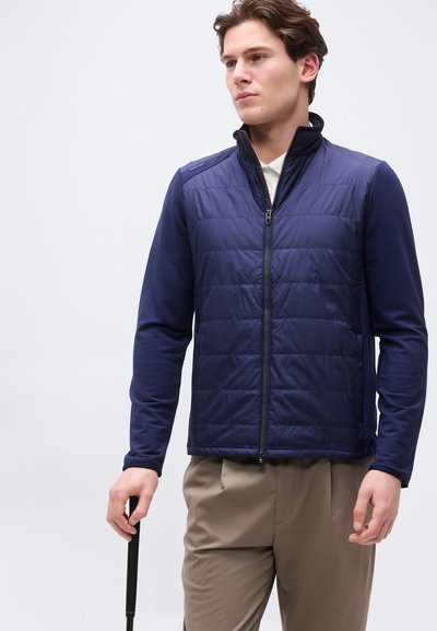 Νεαρός άντρας με κοντά καστανά μαλλιά, φορ ώντας μπουφάν με φερμουάρ από μπλε καπιτονέ ύφασμα πάνω από λευκό πουκάμισο, κρατώντας μαύρη λαβή γκολφ, κοιτάζοντας προς τα δεξιά.