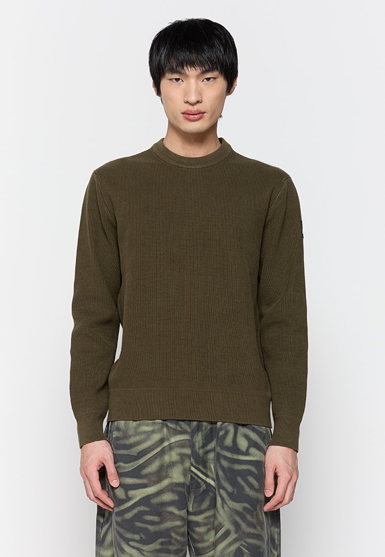Maglione a coste verde oliva con scollatura tonda e maniche lunghe, caratterizzato da una vita aderente e cuciture dettagliate. Indossato con pantaloni con fantasia.
